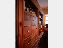 Ein rustikales Holzregal mit mehreren kleinen Schubladen mit Metallgriffen, Flaschen und Gläsern auf den oberen Regalböden, platziert in einem Raum mit warmem Licht und einem Fenster im Hintergrund.