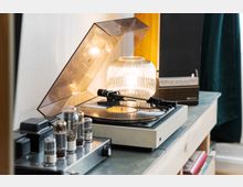 Ein Retro-Schallplattenspieler mit transparentem Deckel steht auf einem Holztisch, daneben eine gläserne Tischlampe und ein Röhrenverstärker. Im Hintergrund sind ein kleiner Lautsprecher und ein Vorhang sichtbar.