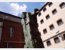 Skulptur im Vordergrund aus Bronze mit rauer Oberfläche, im Hintergrund zwei Gebäudefassaden, eine aus rotem Backstein und eine verputzte weiße Wand mit vergitterten Fenstern. Der Himmel ist teilweise sichtbar, mit blauen Abschnitten und weißen Wolken.
