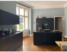 Moderne Küche mit schwarzen Einbaumöbeln und einer Kücheninsel in der Mitte, versehen mit einer hellen Holzparkettboden. Große Fenster mit Blick nach draußen sorgen für viel Tageslicht, und im Hintergrund steht ein Buddha-Dekor auf einem Schrank.