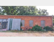 Backsteinbau mit roten Ziegeln und einer grauen Holztür, gekennzeichnet durch ein Schild mit der Aufschrift "Hofladen". Die Fassade ist mit drei Glasbausteinfenstern, einem alten hölzernen Wagenrad und Umrankungen von Pflanzen geschmückt; im Vordergrund befindet sich ein unbefestigter Weg.