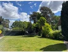 Garten mit Rasenfläche, verschiedenen Bäumen und Sträuchern sowie einem Trampolin und einer Kinderschaukel im Hintergrund. Im oberen Bildbereich sind Wolken und blauer Himmel zu sehen, während rechts ein Haus mit Schrägdach und Hecken sichtbar ist.