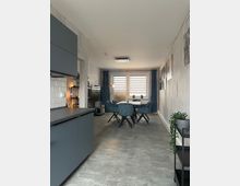 Wohnbereich einer modernen Wohnung mit offener Küche und Essbereich. Graue Küchenmöbel, ein Tisch mit gepolsterten Stühlen, bodentiefe Fenster mit halbtransparenten Rollos sowie moderne Dekorationen und Wandbilder prägen den Raum.