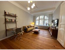 Ein wohnliches Wohnzimmer mit Holzdielen und großen Fensterbögen, durch die Bäume sichtbar sind. Der Raum ist mit einem gelben Sofa, einem Couchtisch, einem Schreibtisch mit Bücherregal und einem Sideboard mit Dekoration ausgestattet, beleuchtet von einer modernen Deckenlampe und einer Stehlampe.