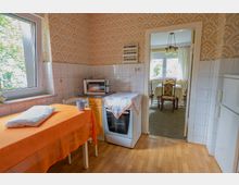 Blick in eine kleine Küche mit großem Fenster, gemusterten Tapeten und halbhoch gefliesten Wänden, mit Tisch, Herd, Mikrowelle und Kühlschrank. Durch eine offene Tür ist ein angrenzendes Esszimmer mit Tisch, Stühlen und Hängelampe zu sehen.