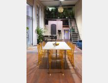 Ein moderner Raum mit hohen Decken und einer Zwischengeschoss-Ebene, die über eine Metalltreppe erreichbar ist. Im Vordergrund steht ein Esstisch mit transparenten gelben Stühlen, umgeben von Pflanzen und großen Fenstern mit weißen Vorhängen.