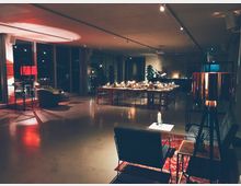 Großer, modern eingerichteter Raum mit festlich gedeckten Tischen im Hintergrund, beleuchtet von Kerzen und warmem Licht. Im Vordergrund befinden sich eine Sitzecke mit einem roten Teppich, zwei Sesseln, einem Beistelltisch und einer großen Stehlampe.