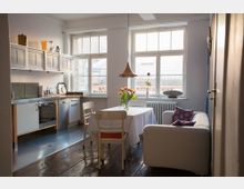 Helle Küche mit Holzboden, ausgestattet mit modernen weißen Schränken, einem Herd und einer Spüle aus Metall. Im Raum steht ein Esstisch mit Tischdecke, Vase mit Tulpen, sowie ein Sofa vor großen Fenstern mit Blick auf Dächer.