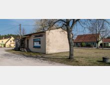 Ein kleiner Dorfladen mit der Aufschrift „Lebensmittel“ in einem beigen Gebäude; davor ein Baum und eine unbefestigte Straße. Im Hintergrund sind Wohnhäuser mit roten Ziegeldächern, grün gestrichene Fassaden und ein Baum zu sehen.