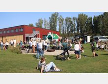 Ein Außenbereich mit einer großen Rasenfläche, auf der sich viele Menschen versammelt haben, darunter eine Blaskapelle. Im Hintergrund steht ein modernes, rot-braunes Gebäude mit Holz- und Metallakzenten sowie eine Hüpfburg in Form einer Burg.