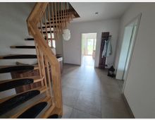 Heller Eingangsbereich eines Wohnhauses mit großer, grauer Fliesenbodenfläche und einer offenen Holztreppe mit Geländer. Rechts stehen eine Garderobe und ein großer Spiegel; durch eine Türöffnung ist ein weiterer Raum mit Blick nach draußen zu sehen.