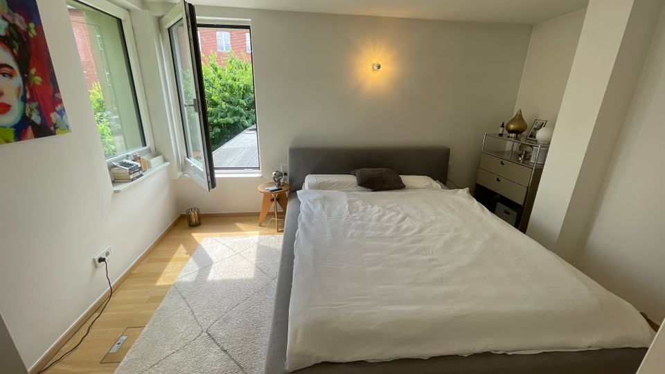 Helles, modern eingerichtetes Schlafzimmer mit Doppelbett, hellem Teppich und Holzfußboden. Links sorgen große, teils geöffnete Fenster für Tageslicht und Blick ins Grüne; rechts stehen eine Kommode und eine Wandlampe an einer schrägen Wandnische.