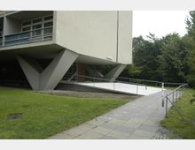 Ein modernes Gebäude mit V-förmigen Betonstützen, die den oberen Teil tragen, sowie einer geneigten Rampe mit Geländer für den Zugang. Im Hintergrund befinden sich Bäume und eine Grünfläche, während eine gepflasterte Fläche den Vordergrund einnimmt.