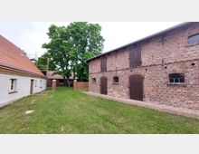 Ein Innenhof mit einer ziegelroten Scheune auf der rechten Seite und einem weiß verputzten Gebäude mit roten Ziegeldächern auf der linken Seite. Im Hintergrund befinden sich Bäume und eine Ziegelmauer.