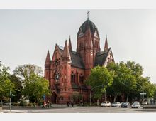 Neugotische Backsteinkirche mit mehreren hohen Türmen und einer zentralen Kuppel mit Uhr, umgeben von Bäumen und einer Straßenkreuzung mit Fußgängern, Fahrrädern und parkenden Autos.