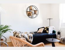 Modern eingerichtetes Wohnzimmer oder Gästezimmer mit hellem Holzboden, einem Tagesbett mit schwarzer Bettdecke und dekorativen Kissen, einer runden Wandregalstruktur aus Metall und Holz, einer Stehlampe und einer Zimmerpflanze. Auf der rechten Seite fällt Tageslicht durch ein weißes Fenster mit Vorhängen.