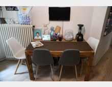 Ein kleiner Essbereich mit einem Holztisch und einer Tischdecke, umgeben von drei Stühlen (zwei graue, ein weißer). Auf dem Tisch befinden sich Zeitschriften, ein Kochbuch, ein Tablet, ein Luftbefeuchter und ein Mixer. Im Hintergrund sind eine weiße Wand, ein Heizkörper und ein an der Wand montierter schwarzer Fernseher zu sehen.