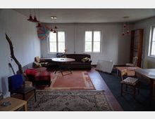 Ein schlicht eingerichteter Wohnraum mit weißen Wänden, zwei Fenstern und mehreren Möbelstücken, darunter ein Sofa, Stühle, Tische und Regale. Farbige Teppiche bedecken den Boden, und an der Decke hängen rote Lampen; an der Wand ist ein buntes Kunstwerk angebracht.