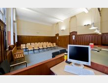 Ein Sitzungssaal mit Holzwänden und dekorativem Wandpaneel, bestückt mit einem Halbkreis aus Holzstühlen. Im Vordergrund befindet sich ein Schreibtisch mit einem Computermonitor und einer Tastatur, während helle Hängelampen die Szene beleuchten.