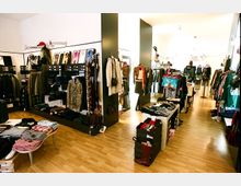 Ein heller Laden mit Holzboden, in dem Kleidung, Schuhe und Accessoires ausgestellt sind. Die Regale und Kleiderstangen sind entlang der Wände und in der Mitte des Raumes in Reihen angeordnet.