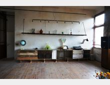 Holzfußboden in einem Raum mit einer rustikalen Anrichte aus alten Holzschubladen und -türen. Darüber hängt ein schlichtes Regal mit verschiedenen dekorativen Objekten, darunter Vasen und eine Retro-Waage; daneben eine Tischlampe, und Tageslicht strömt durch ein großes Fenster mit Jalousien ein.