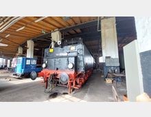 Eine historische schwarze Dampflokomotive mit roten Details steht in einer großen Werkstatthalle mit Holzdach und weißen gemauerten Säulen. Im Hintergrund sind weitere Schienenfahrzeuge und Werkbänke zu sehen.