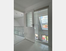 Innenraum eines modernen, hellen Raumes mit weißen Wänden und Boden. Eine Glastür links führt zu einem Raum mit Glasgeländer, während eine Balkontür rechts Ausblick auf eine städtische Umgebung mit blauen Himmel und Wolken bietet.