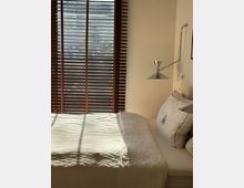 Minimalist bedroom with a neatly made bed in white bedding beside a large window fitted with dark horizontal blinds. Sunlight casts striped shadows across the bed, and a wall-mounted metal reading lamp is positioned above the pillows.