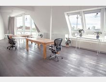 Heller Büroraum mit großen Dachfenstern, durch die eine Stadtkulisse sichtbar ist. Der Raum enthält Holztische, moderne Bürostühle, weiße Schreibtische und Zimmerpflanzen, mit hellen Wänden und einem dunklen Laminatboden.