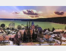 Luftaufnahme eines kleinen Dorfes mit einer markanten Backsteinkirche im Zentrum, umgeben von Fachwerkhäusern und Bäumen. Im Hintergrund erstrecken sich weite grüne Felder und ein Wald unter einem Himmel mit ziehenden Wolken.