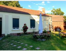 Ein gepflegter Garten mit einer Rasenfläche und mehreren Sitzgruppen aus Holz und Metall, zentral ein Sonnenschirm. Im Hintergrund steht ein Gebäude mit gelber Fassade, schwarzen Rollläden und roten Ziegeln, daneben ein weiteres Gebäude aus rotem Backstein.