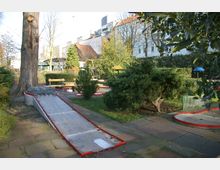 Eine Minigolfanlage in einem parkähnlichen Außenbereich mit mehreren Bahnen, umgeben von Bäumen, Sträuchern und Sitzbänken. Im Hintergrund sind Häuserfassaden sowie begrünte Bereiche zu sehen.