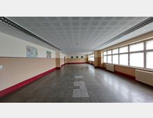 Langer, heller Flur in einem Gebäude mit hellbeigen Wänden, rotem Sockel und großer Fensterfront rechts. An den Wänden hängen bunte Bilder, und die Decke ist mit perforierten Paneelen und langen Leuchtbändern ausgestattet.