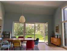 Esszimmer mit einem großen Holztisch und bunten Stühlen, davor eine Glasfront mit Blick auf einen grünen Garten. Rechts steht eine Holzkommode, darüber hängt ein Bild, und eine Designer-Hängelampe ist an der Decke befestigt.