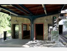 Überdachte Bahnsteigszene mit einer gelben Betonwand, zwei geöffneten Türen mit roten Rahmen und Gitterfenstern. Rechts ein Schienenbereich, ein Signalsystem und ein beschriftetes Glaspaneel; im Hintergrund sind grüne Bäume sichtbar.