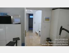 Blick in einen schlichten Innenraum mit weiß gestrichenen Wänden und hellem Holzfußboden, durch eine offene Tür zu einem Treppenaufgang und einem angrenzenden Raum. Links steht ein weißer Schrank, rechts ist ein Staubsauger erkennbar.