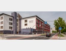 Straßenansicht einer modernen Oberschule mit weißer Fassade, blauen Kacheln und roten Akzenten um die Fenster. Vor dem Gebäude verläuft eine Straße mit Zebrastreifen, Verkehrszeichen und einer Tempo-30-Zone, flankiert von einem Gehweg und Bäumen.