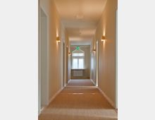 Ein schmaler, beige gestrichener Flur mit Teppichboden, flankiert von symmetrischen Türen auf beiden Seiten. Am Ende des Flurs befindet sich ein Fenster mit einer kleinen Heizung darunter, darüber ein leuchtendes Notausgangsschild.