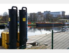 Industrielles Hafengebiet mit zwei großen schwarzen Zylindern und einem gelben Container im Vordergrund, einem Wasserbecken mit Frachtschiff und farbigen Bojen sowie modernen Bürogebäuden und kahlen Bäumen im Hintergrund.