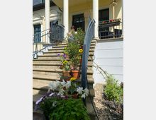 Eine Treppe mit Geländer führt zu einem Gebäude mit gelber Fassade, großen Fenstern und weißen Säulen. Die Treppenstufen sind mit zahlreichen Blumentöpfen in verschiedenen Größen und bunten Blumen dekoriert.