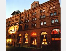 Backsteinfassade eines mehrstöckigen historischen Gebäudes mit großen Rundbogenfenstern im Erdgeschoss, die von innen orange beleuchtet sind. Das Gebäude zeigt klassische industrielle Architekturdetails, und der Himmel im Hintergrund ist im Abendlicht sichtbar.