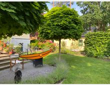 Garten mit Rasenfläche, einem kleinen Baum mit dichtem, kugelförmigem Blätterdach und umliegenden Blumenbeeten. Im Vordergrund eine Terrasse mit einer gelb-orangen Hängematte, einem Stuhl, Blumentöpfen und einer Treppe, die zu einem hellgelben Gebäude führt.