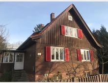 Ein zweistöckiges Holzhaus mit einer Fassade aus braunem Holz und roten Fensterläden. Das Gebäude hat ein steiles Satteldach mit roten Ziegeln, eine kleine Treppe, die zu einer Glastür führt, und ist von Bäumen und einem Holzzaun umgeben.