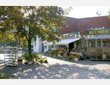 Blumengeschäft mit gelber Aufschrift „Blumen“ auf einem grünen Schild, umgeben von Blumen- und Pflanzenarrangements in Töpfen und Kisten; im Vordergrund ein gepflasterter Platz mit einem einzelnen Baum, dahinter ein Backsteingebäude.
