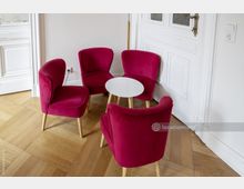 Vier dunkelrote Polsterstühle mit Holzbeinen stehen um einen kleinen weißen runden Tisch auf einem Boden aus hellem Parkett. Im Hintergrund sind eine weiße Wand und eine Tür mit verzierten Kassettenelementen zu sehen.