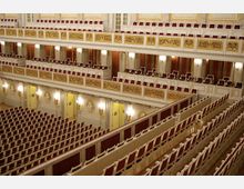 Innenraum eines prunkvollen Konzert- oder Theatersaals mit mehreren Rängen. Die Sitzplätze sind mit roten Polstern versehen, und die Balkone sind mit weißen und goldenen Verzierungen sowie Wandreliefs und goldenen Wandlampen gestaltet.