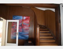 Holzgetäfelter Treppenbereich mit einer geschwungenen Holztreppe und einem großen abstrakten Gemälde in Rot- und Blautönen an der Wand. Links steht eine geöffnete weiße Tür mit einer strukturierten Glaseinlage.