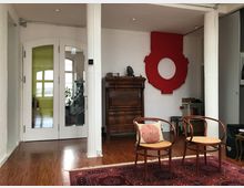 Wohnraum mit Holzboden, einer roten Wanddekoration in Form eines Kreises, einem antiken Holzschrank und zwei Holzstühlen mit geflochtenen Sitzflächen. Im Hintergrund sind eine Glastür und ein Raum mit großen Fenstern sichtbar.