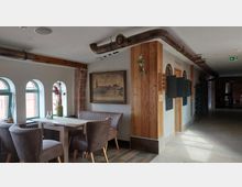 Gemütlicher Innenraum mit einem kleinen Sitzbereich, bestehend aus gepolsterten Sesseln und einer Bank an einem Holztisch, vor drei gewölbten Fenstern mit grünen Rahmen. An der Wand hängt ein Gemälde, und sichtbare Backstein- sowie Holzelemente verleihen der Szenerie ein rustikales Ambiente; ein Rohrsystem verläuft entlang der Decke.