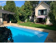 Privater Garten mit einem rechteckigen Swimmingpool im Vordergrund, umgeben von hellen Steinplatten. Im Hintergrund ein zweistöckiges Haus mit heller Fassade und einem Balkon mit Glas-Schiebetür, sowie eine Terrasse mit Sitzmöbeln und Sonnenschirm. Rundherum sind Bäume, Büsche und ein gepflegter Rasen.